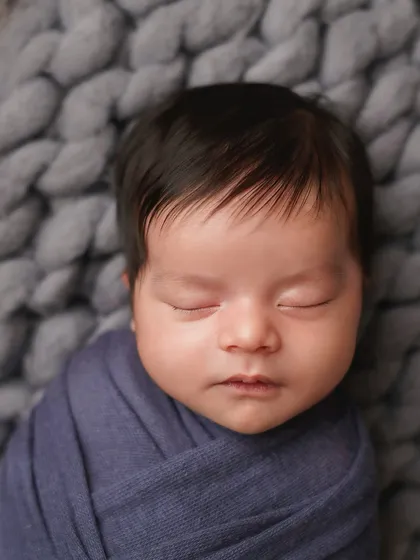 A close-up on the face of the newborn in the blue wrap, highlighting his peaceful expression and beautiful dark hair.