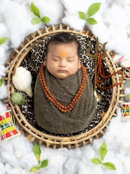 A serene overhead shot of a newborn styled as Lord Shiva. The setup includes a trishul, damaru, and shankha, all nestled in soft cotton clouds.