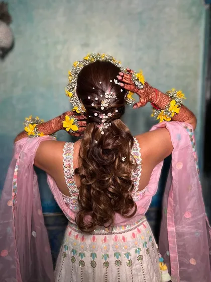 A back view of this pretty Haldi hairstyle. The hair is styled in a curly ponytail, adorned with pearls and a fresh yellow floral crown.