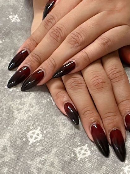 A dark and dramatic ombre on long stiletto nails, transitioning from a deep red or maroon base to a solid black tip.
