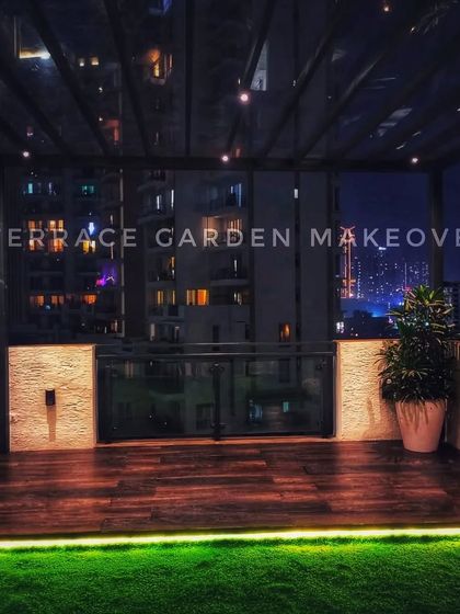 A sleek and modern balcony design featuring a glass roof, wooden deck flooring, and an artificial grass border. The strip lighting under the deck edge adds a sophisticated touch for the evening.