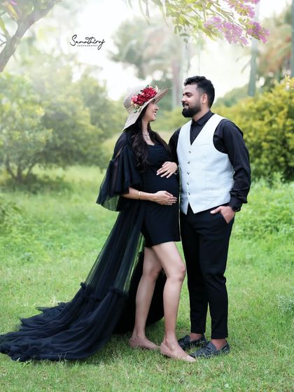 A stylish outdoor couple portrait. The wife, in a black dress and hat, shares a loving gaze with her husband, creating a chic and romantic image in a natural setting.