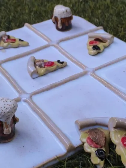 A close-up of a whimsical, handmade ceramic tic-tac-toe set with pizza slices and beer mugs as game pieces. We encourage you to bring your most creative and playful ideas to life in our studio.