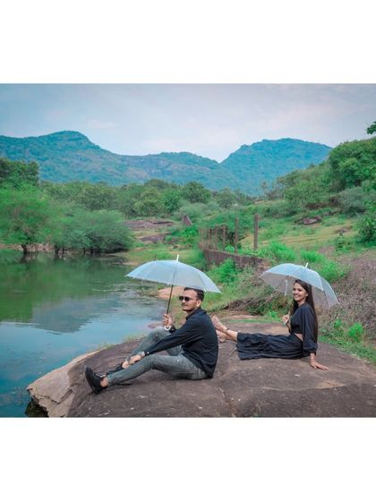 A relaxed pre-wedding scene where a couple sits on a large rock by a lake, holding umbrellas and enjoying the beautiful mountain view.