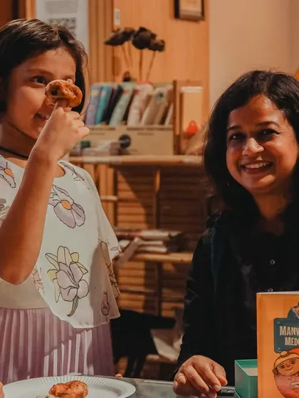 The pure joy of a book event. A young reader playfully enjoys a medu vada during the launch of 'Manvinder's Medu Vada', with the author smiling beside her.