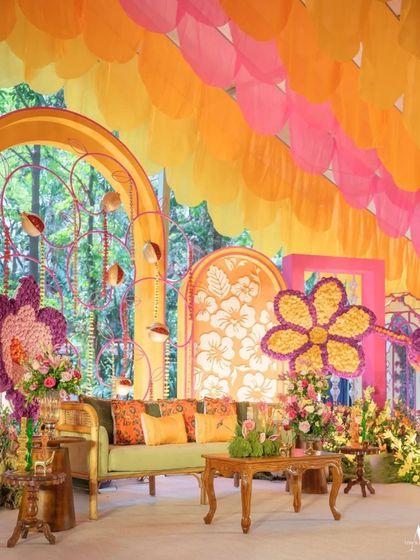 A vibrant and whimsical mehendi stage. The decor features a green sofa, large floral cutouts, and a backdrop of orange and pink arches, creating a playful and artistic setting.