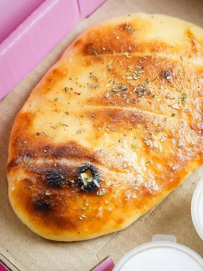 A close-up of a freshly baked garlic bread or flatbread in a pizza box. The focus on the golden-brown crust and herbs highlights its simple, comforting appeal.