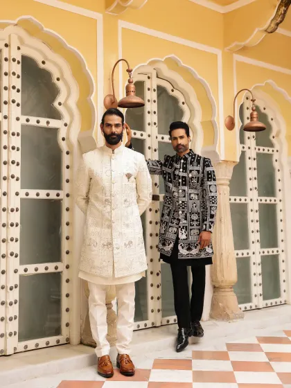 A study in contrasts, this image features two of our signature ceremonial looks. One is a classic off-white embroidered sherwani, and the other is a contemporary black and white patterned bandhgala, showing our range from traditional to modern.