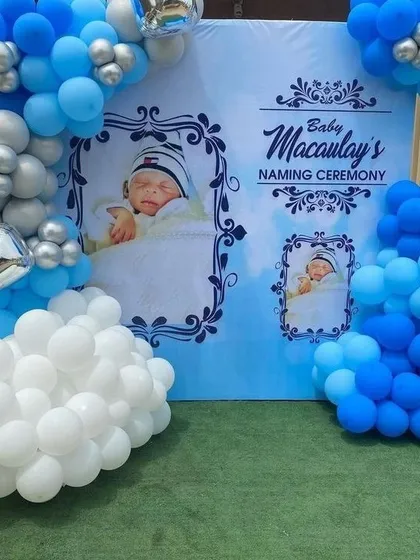 A personalized backdrop for a baby's naming ceremony. This design features a custom printed banner with the baby's photos, framed by a cloud-like arrangement of white, blue, and silver balloons.