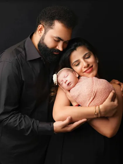 A classic family portrait against a dark background. The parents' loving gaze on their newborn creates a timeless and emotional image.
