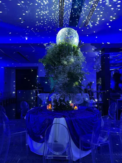 A beautifully set table at the blue-themed birthday party. A large centerpiece with a disco ball and baby's breath sits on a table draped in velvet, under a ceiling of twinkling lights that look like a galaxy.