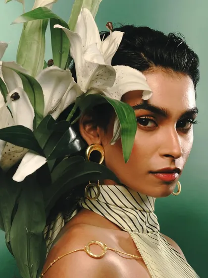 A beauty and jewelry photograph for Batoki.art, where the model's profile is framed by white lilies against a soft green backdrop. The focus is on the elegant gold earrings and the serene, powerful mood of the composition.