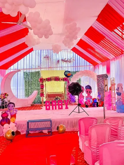 An indoor view of a baby shower pandal, showing the stage with a moon prop, swing, and balloon decorations.