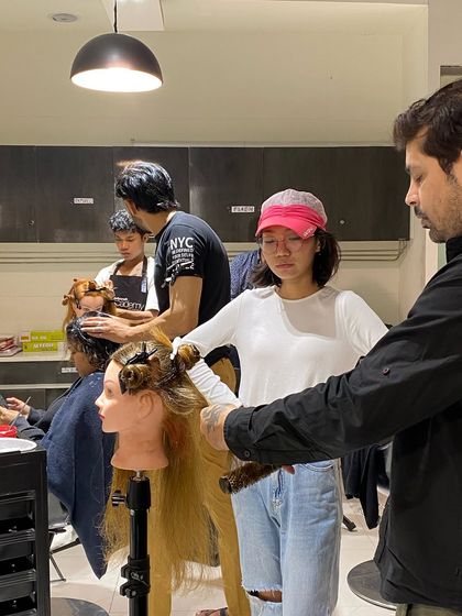 An instructor guides a student through a styling technique on a mannequin, ensuring they understand the hand positioning and tool manipulation required.