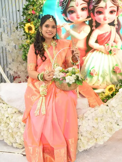 A smiling mom-to-be at her baby shower, holding a floral basket and wearing a matching jewellery set. The peach and white flowers create a soft and elegant look.