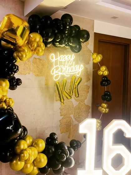 A stylish sweet sixteen birthday setup. The decor features a black and gold balloon ring, a 'Happy Birthday' neon sign, and large light up marquee numbers for the age '16'.