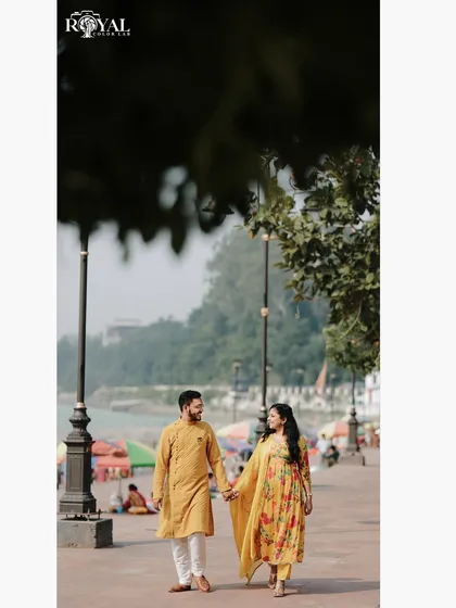 A full-length shot of a couple walking hand-in-hand along the river promenade, framed by lush green trees.