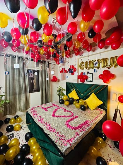 A romantic bedroom setup with red, black, and gold balloons. The bed is covered in petals spelling "I <3 U", and balloons with photos hang from the ceiling.
