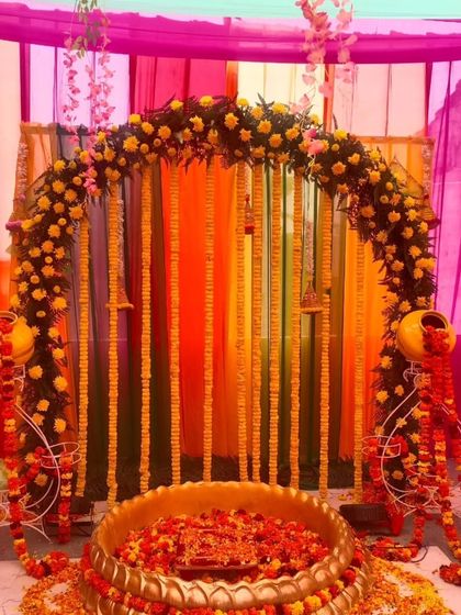 A traditional Haldi ceremony setup with a floral arch and a large brass urli. The decor uses marigold garlands and decorative matkas on stands, perfect for a home-based function.