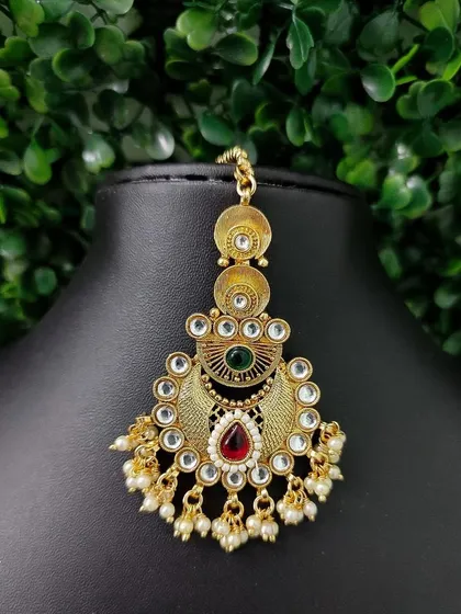 A traditional gold-finish maang tikka with red and green stones.