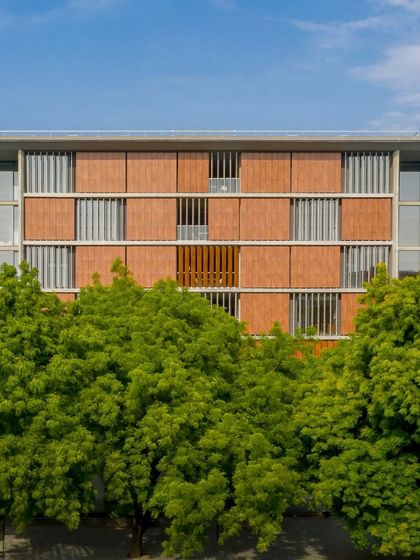The facade of the School of Arts and Sciences at Ahmedabad University, seen through a frame of lush green trees. The building's material palette of concrete and terracotta complements the natural surroundings.