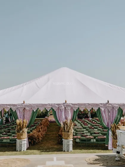 An outdoor wedding tent with a jungle-inspired, floral-free theme. The entrance is flanked by pampas grass, leading into a space filled with green and pastel drapes and seating.