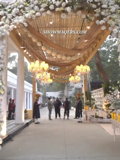 A magnificent floral entrance arch for a daytime celebration. The archway is covered in white flowers, leading into a covered walkway with draped fabric and hanging chandeliers.