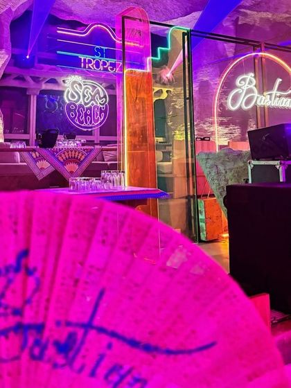 The party in full swing, with neon signs for 'St Tropez,' 'Sex on the Beach,' and 'Bastian' lighting up the cave-like venue.