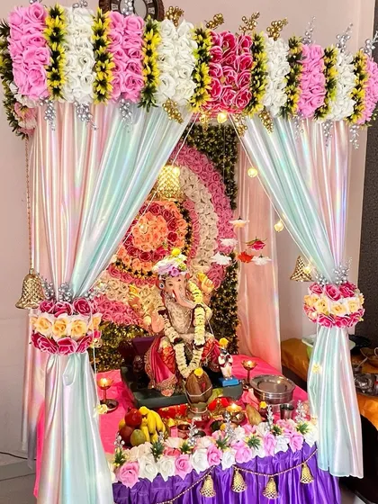 A vibrant and colorful Ganpati mandap featuring a multi-layered floral backdrop in concentric circles. The iridescent fabric and hanging bells add a playful and festive touch to the celebration.