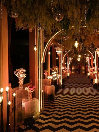 An entrance that whispers tales of elegance and love. For this Sangeet, we created a walkway with a chevron-patterned floor, lined with glowing floral pedestals and framed by modern arches.