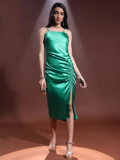 This emerald green satin dress is styled for a formal event. The side slit and ruched details are highlighted by the focused lighting, and the look is completed with metallic heels and a simple necklace.
