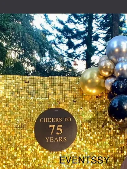 A glamorous "Cheers to 75 Years" photo backdrop. The shimmering gold sequin wall provides a perfect background for photos, complemented by a classic black, gold, and silver balloon garland.
