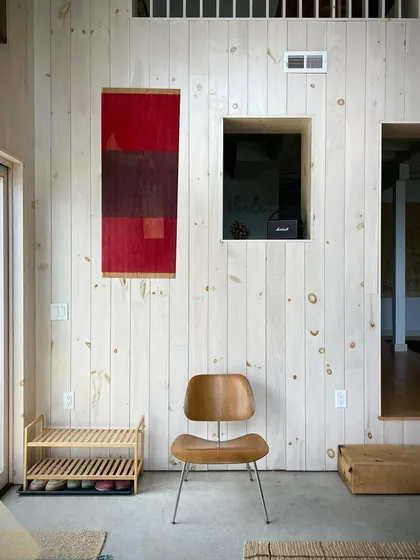 A quiet corner inside the Crooked Lake house, featuring whitewashed pine walls, a classic Eames chair, and a textile piece by artist Malene Bach. The interior design emphasizes simplicity, craftsmanship, and natural materials.