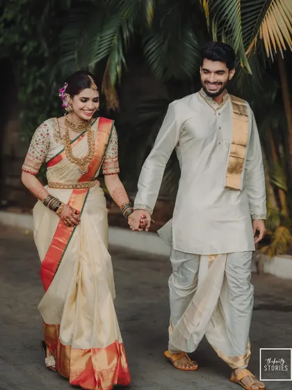A classic South Indian couple portrait, showcasing the bride's elegant off-white and red silk saree and the groom's traditional veshti. Their happy smiles as they walk hand-in-hand reflect the joy of their wedding day.