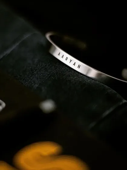 A silver cuff with the name 'AARYAN' resting on a dark fabric, showcasing the crispness of the permanent laser engraving.