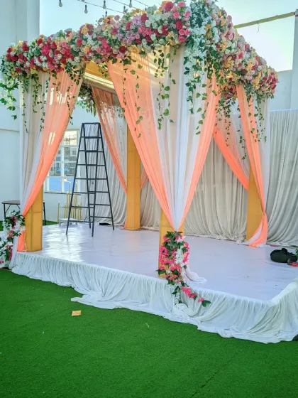 A four-pillar mandap setup on a raised stage, decorated with peach and white drapes and a crown of pastel-colored flowers.