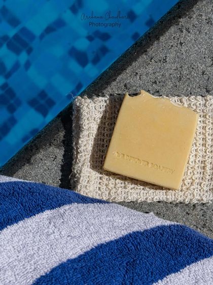 A minimalist composition featuring a single yellow bar of soap on a washcloth. The diagonal lines of the pool edge and towel create a strong visual path to the product.