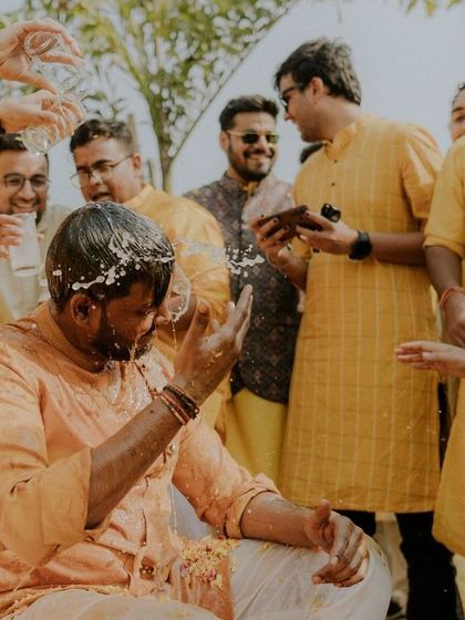 The groom reacts with a laugh as he wipes turmeric and flowers from his face, a genuine, unposed reaction.