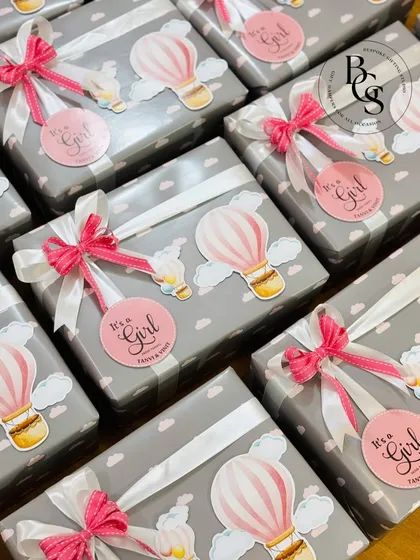 A set of beautifully wrapped baby announcement gifts. The custom paper features clouds and hot air balloons, and each box is finished with a pink "It's a Girl" sticker and a delicate ribbon.