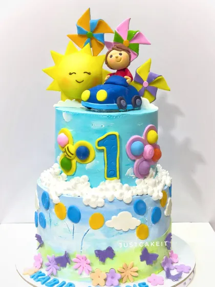 A first birthday cake with a fun, summery theme. The design includes a smiling sun, a little boy in a car, and colorful pinwheels.