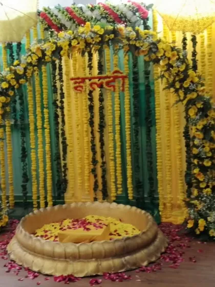 A traditional Haldi setup with a floral arch made of yellow flowers and baby's breath. The large brass urli is filled with petals, set against a backdrop of green and yellow marigold strings.