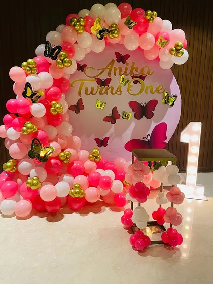 This is a popular butterfly theme decoration I did for a first birthday. It features a dense balloon arch in shades of pink and white, accented with gold and pink butterflies, and a large light-up marquee number '1'.