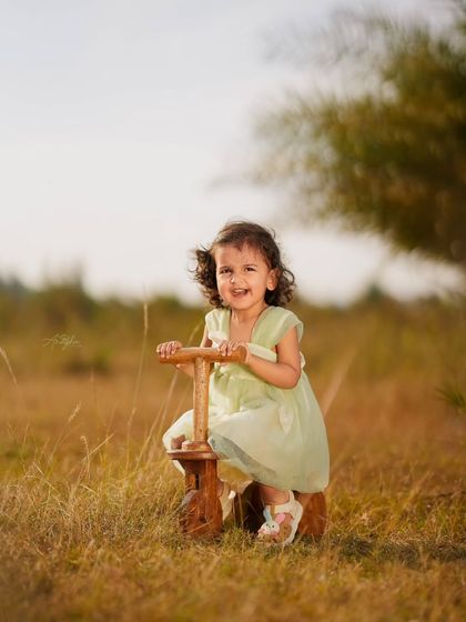 A happy toddler laughs while sitting on a wooden tricycle in a field of tall grass, bathed in beautiful golden hour light.