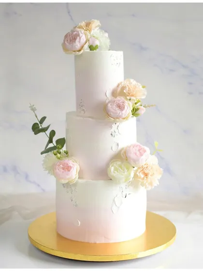 A classic three-tiered wedding cake with a subtle pink ombre effect on the bottom tier. The cake is decorated with silver leaf and clusters of beautiful fresh flowers for a timeless and romantic feel.