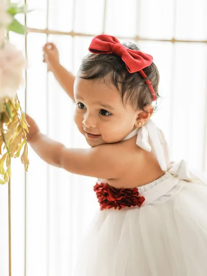 A beautiful, classic portrait of a baby girl. The red bow and floral details on her white dress add a festive touch, perfect for a milestone or holiday-themed photoshoot.