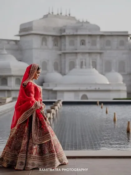 A stunning bridal portrait of the bride in her red lehenga, set against the magnificent white facade of a palace hotel in Jaipur.