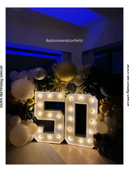 Fifty and fabulous. This 50th birthday setup features bold marquee numbers surrounded by a classic black, white, and gold balloon arch.