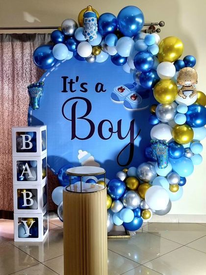 A classic 'It's a Boy' baby shower decoration. The setup includes a large circular backdrop, a blue, white, and silver balloon garland, and 'BABY' blocks.