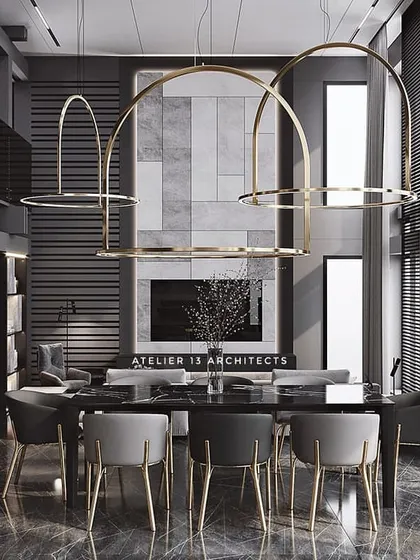 A dining room with a bold, monochromatic design. Large, arched brass chandeliers hang over a black dining table, while grey and black chairs with brass legs continue the theme. The double-height space feels grand and contemporary.