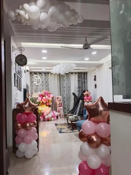 A soft and sweet 'Welcome Baby' decoration inside a home. The setup includes balloon columns, clouds of white balloons on the ceiling, and a cozy canopy area.